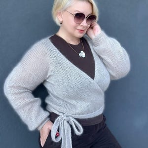 May include: A light gray, wrap-style sweater with long sleeves and a tie closure. The sweater is made of a soft, fuzzy material. The model is wearing a brown top and black pants. The background is a solid, dark blue color.