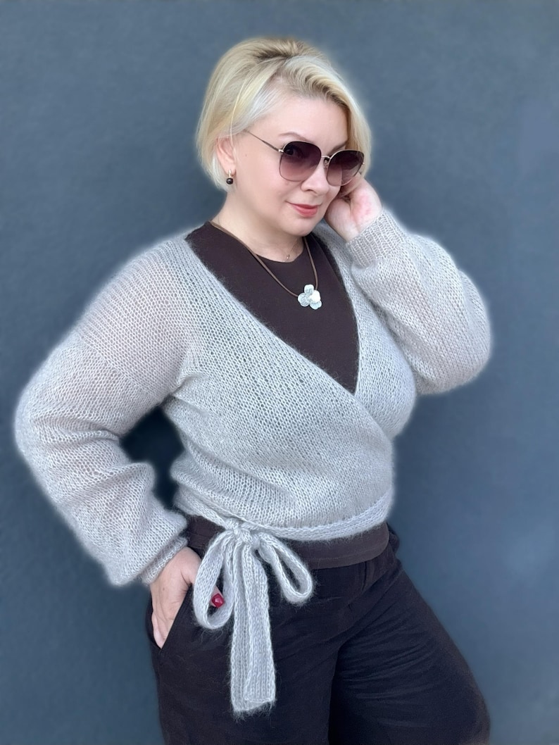 May include: A light gray, wrap-style sweater with long sleeves and a tie closure. The sweater is made of a soft, fuzzy material. The model is wearing a brown top and black pants. The background is a solid, dark blue color.