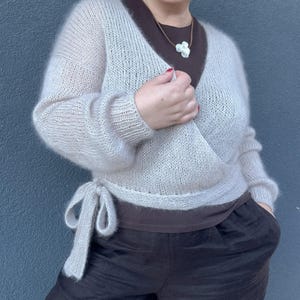 May include: A light beige wrap sweater with long sleeves and a tie closure. The sweater is paired with a brown top and dark brown pants. The outfit is completed with a white flower pendant necklace.