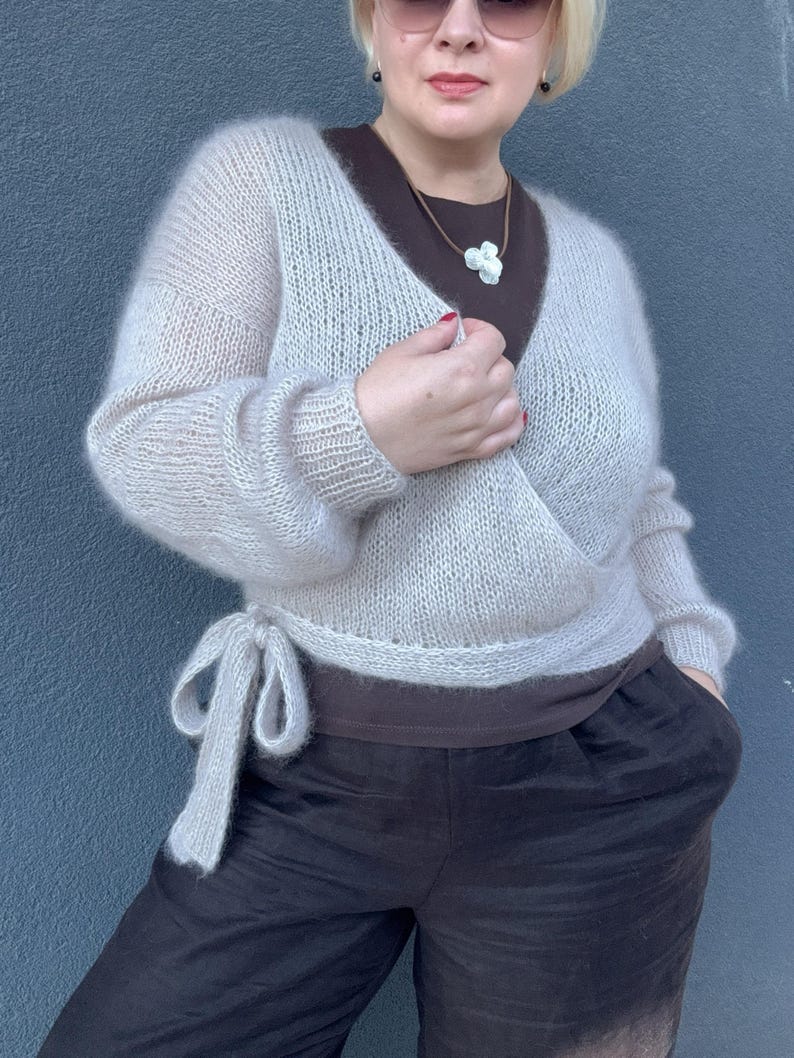 May include: A light beige wrap sweater with long sleeves and a tie closure. The sweater is paired with a brown top and dark brown pants. The outfit is completed with a white flower pendant necklace.