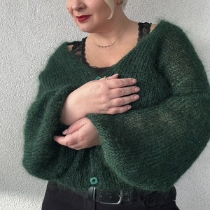 Puede incluir: Un c&aacute;rdigan de color verde oscuro y peludo con cierre de bot&oacute;n. El c&aacute;rdigan se lleva sobre un top de encaje negro y pantalones negros.