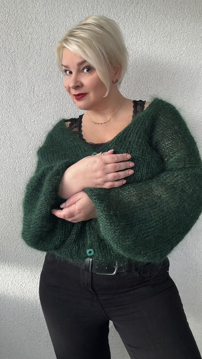 Puede incluir: Un c&aacute;rdigan de color verde oscuro y peludo con cierre de bot&oacute;n. El c&aacute;rdigan se lleva sobre un top de encaje negro y pantalones negros.