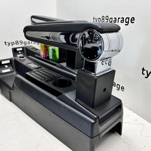 May include: A black and chrome car console with a padded armrest. The console features a chrome gear shift and cup holders. The text "tvp89garage" and "typ89" are visible on the background.