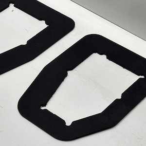 Peut inclure: Deux joints en caoutchouc noir avec une découpe rectangulaire au centre. Les joints sont sur une surface blanche.