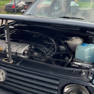 Könnte beinhalten: Ein schwarzer Volkswagen Golf mit geöffneter Motorhaube, die den Motor und einen durchsichtigen Plastikbehälter freilegt. Das Auto hat einen schwarzen Kühlergrill mit dem Volkswagen-Logo und eine schwarze Stoßstange.