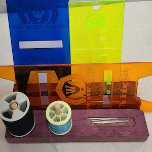 May include: A purple wooden sewing tool with two spools of thread, needles, and two clear plastic rulers. The rulers are labeled "Two Woolf Designs" and "The Binding Buddy".