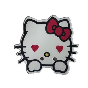 Puede incluir: Un diseño blanco de Hello Kitty con un lazo rojo y ojos en forma de corazón. El contorno es negro y el diseño tiene dos pequeños agujeros. El diseño general es un personaje lindo y reconocible.