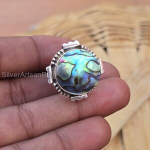 Pode incluir: Um anel de prata com uma incrustação redonda de concha de abalone. A concha tem cores iridescentes de azul, verde e roxo.