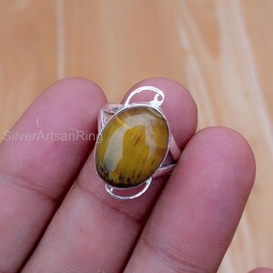 May include: A silver ring featuring an oval-shaped tiger's eye gemstone. The gemstone is a mix of golden-brown and yellow hues. The ring has a simple, elegant design with a decorative loop above the gemstone. The text "SilverArtsanRing" is visible.