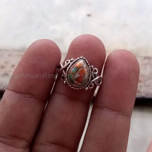 May include: A silver ring featuring a teardrop-shaped stone with a mix of orange, teal, and white colors. The ring has an ornate, twisted metal design around the stone and along the band. The ring is shown on a person's finger.