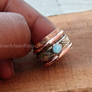 May include: A silver and copper spinning ring with a light blue gemstone. The ring has a textured design and is worn on a finger.