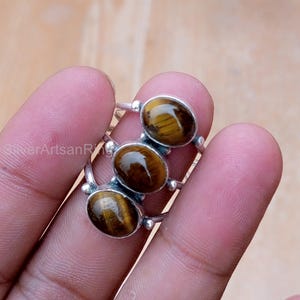 May include: A silver ring with three oval tiger's eye stones. The stones are a warm brown color with golden streaks. The ring is held by a person.