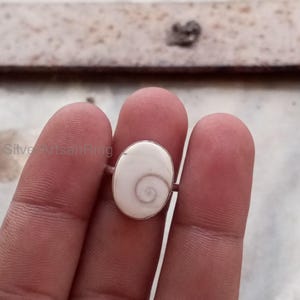 Könnte beinhalten: Ein Silberring mit einem ovalen, cremefarbenen Stein mit einem Spiralmuster. Der Ring wird zwischen Fingern gehalten, wodurch der polierte Stein und das schlichte Band hervorgehoben werden. Das Design des Rings ist minimalistisch und elegant.