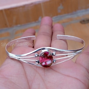 Puede incluir: Una pulsera de plata con un centro de piedra ovalada rosa y blanca. La pulsera tiene un diseño delicado y abierto con tres líneas paralelas a cada lado de la piedra. La piedra tiene un patrón natural único.