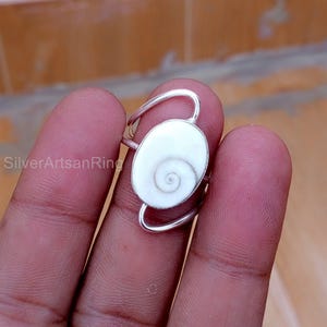 Puede incluir: Un anillo de plata con una piedra ovalada blanca con un diseño en espiral. La banda del anillo se curva alrededor de la piedra, creando un aspecto único. El anillo se muestra en un dedo.