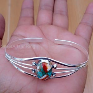 Puede incluir: Una pulsera de plata con una piedra central ovalada en colores rojo, turquesa y blanco. La pulsera tiene un diseño de tres hilos a cada lado de la piedra. Se muestra en una mano.
