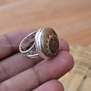 Pode incluir: Anel de prata com uma gema de amonite marrom engastada em uma moldura de prata. A banda do anel é de prata e tem um design decorativo.
