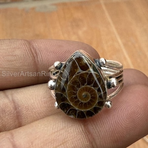 Może przedstawiać: Srebrny pierścionek z brązowo-beżowym kamieniem kopalnym w kształcie łzy. Kamień ma spiralny wzór. Pierścionek ma wielopasmowy design z małymi srebrnymi kulkowymi akcentami. Widoczny jest tekst "SilverArtsanRing".