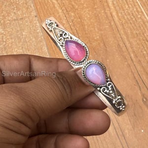 Puede incluir: Pulsera de plata con dos gemas en forma de lágrima en tonos rosa y lavanda. La pulsera tiene un diseño ornamentado con detalles decorativos plateados. El texto "SilverArtsanRing" es visible.