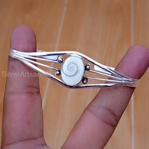 Puede incluir: Una pulsera de puño plateada con un elemento central blanco en forma de espiral. La pulsera tiene un diseño delicado y abierto con tres barras paralelas a cada lado de la pieza central y pequeños acentos redondos.