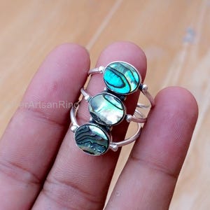 Puede incluir: Tres anillos de plata, cada uno con una concha de abulón ovalada. Las conchas muestran iridiscencias azules, verdes y blancas. Los anillos son sostenidos por los dedos de una persona, con una superficie de madera en el fondo.