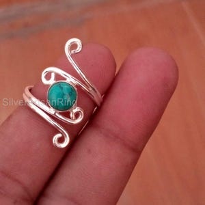 May include: A silver ring with a turquoise stone. The ring has a unique design with swirling, openwork details. The turquoise stone is round and set in the center of the ring. The ring is shown on a finger.