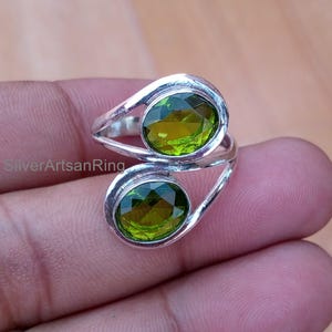Puede incluir: Un anillo de plata con dos piedras de peridoto de corte ovalado. Las piedras de color verde brillante están engastadas en un diseño contemporáneo. El anillo se muestra en una mano. Se ve el nombre de la marca "SilverArtsanRing".