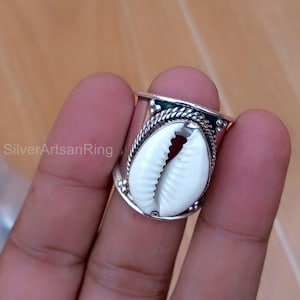 Peut inclure: Bague en argent ornée d'un coquillage cauri blanc. La bague a une large bande avec des détails décoratifs autour de la coquille. Le texte "SilverArtsanRing" est visible sur le côté gauche de l'image.