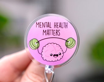 Pode incluir: Um porta-crachá de plástico rosa e transparente com um cérebro de desenho animado levantando pesos. O texto no porta-crachá diz "Mental Health Matters".