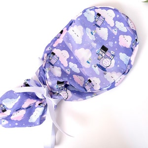 May include: A purple scrub hat with a white ribbon tie. The hat has a repeating pattern of white clouds with pink accents and a cartoon medical cart with the text "Thank you for being a nurse".