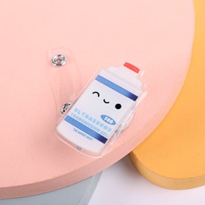 May include: A clear plastic badge holder with a cartoon image of a bottle of ultrasound transmission gel. The bottle has a red lid and a smiling face. The text on the bottle reads "Ultrasound Transmission Gel 100ml The Nudge Spot".