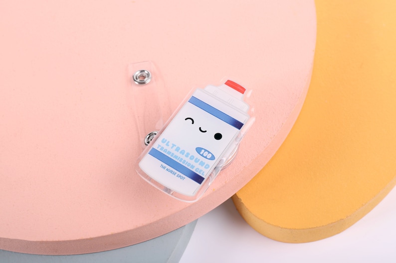 May include: A clear plastic badge holder with a cartoon image of a bottle of ultrasound transmission gel. The bottle has a red lid and a smiling face. The text on the bottle reads "Ultrasound Transmission Gel 100ml The Nudge Spot".