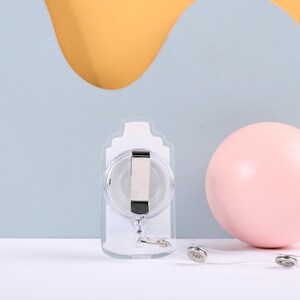 May include: A clear plastic retractable badge holder with a silver clip and a silver chain. The holder is on a white surface with a pink ball and a yellow and blue background.