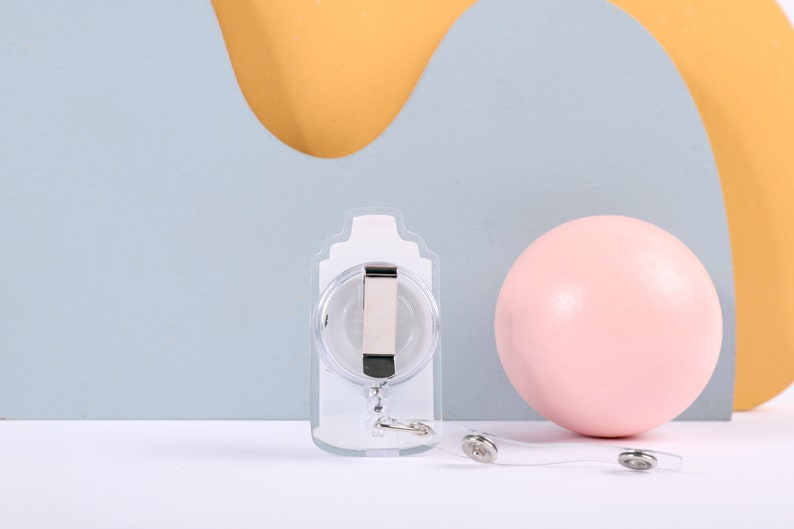 May include: A clear plastic retractable badge holder with a silver clip and a silver chain. The holder is on a white surface with a pink ball and a yellow and blue background.