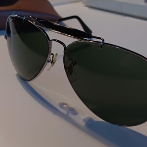 May include: Aviator-style sunglasses with dark green lenses and a silver metal frame. The sunglasses have a top bar and thin temples. A brown leather case is visible in the background.