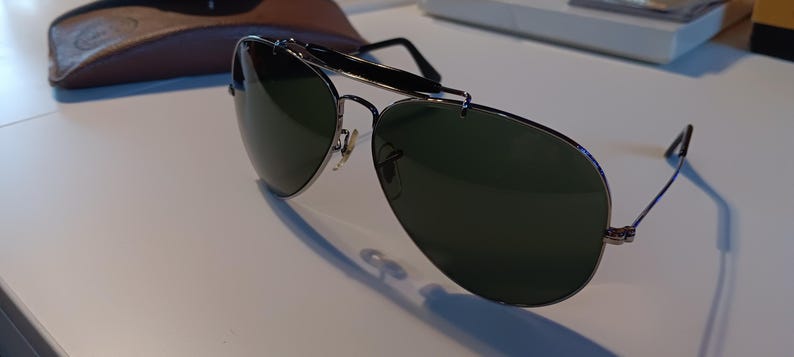 May include: Aviator-style sunglasses with dark green lenses and a silver metal frame. The sunglasses have a top bar and thin temples. A brown leather case is visible in the background.