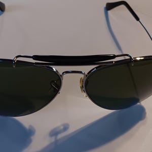 May include: Aviator-style sunglasses with dark green lenses and a silver frame. The sunglasses have a black bridge and temples. The Ray-Ban logo is visible on the left lens. The sunglasses are on a white surface.