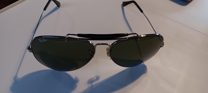 May include: Aviator-style sunglasses with dark green lenses and a silver frame. The sunglasses have a black bridge and temples. The Ray-Ban logo is visible on the left lens. The sunglasses are on a white surface.