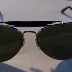 May include: Aviator-style sunglasses with dark green lenses and a black brow bar. The sunglasses have a silver metal frame and black temples. The lenses are teardrop-shaped, and the bridge has a double bar design.