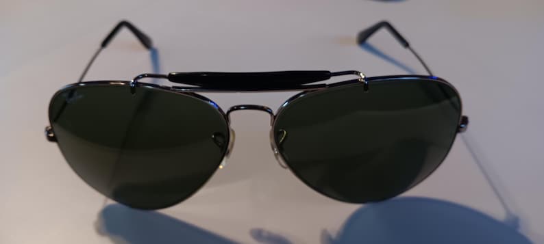 May include: Aviator-style sunglasses with dark green lenses and a black brow bar. The sunglasses have a silver metal frame and black temples. The lenses are teardrop-shaped, and the bridge has a double bar design.