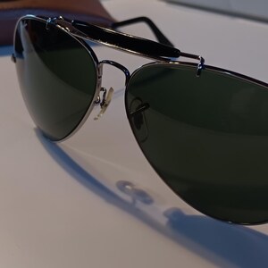 May include: Aviator-style sunglasses with dark green lenses and a silver metal frame. The sunglasses have a double bridge and a black bar across the top. A brown leather case is visible in the background.