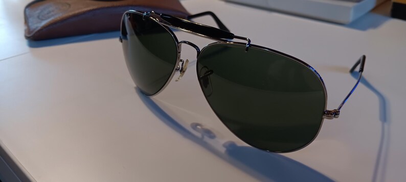 May include: Aviator-style sunglasses with dark green lenses and a silver metal frame. The sunglasses have a double bridge and a black bar across the top. A brown leather case is visible in the background.
