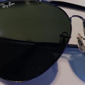 May include: Aviator-style sunglasses with dark green lenses and a silver metal frame. The Ray-Ban logo is visible on the upper left lens. The sunglasses have a double bridge and curved temples.