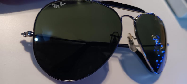 May include: Aviator-style sunglasses with dark green lenses and a silver metal frame. The Ray-Ban logo is visible on the upper left lens. The sunglasses have a double bridge and curved temples.