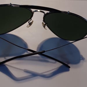 May include: Aviator-style sunglasses with a black metal frame and dark green lenses. The sunglasses are resting on a white surface, casting a shadow. The design features a double bridge and curved temples.