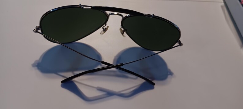 May include: Aviator-style sunglasses with a black metal frame and dark green lenses. The sunglasses are resting on a white surface, casting a shadow. The design features a double bridge and curved temples.