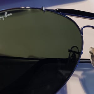 May include: Aviator-style sunglasses with dark green lenses and a silver frame. The Ray-Ban logo is visible on the upper left lens. The sunglasses have a double bridge and nose pads.