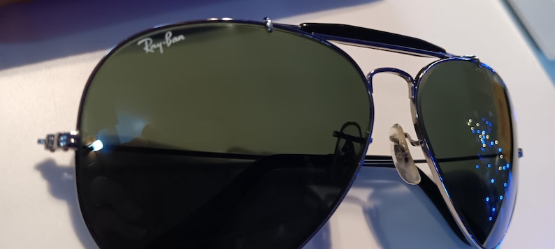 May include: Aviator-style sunglasses with dark green lenses and a silver frame. The Ray-Ban logo is visible on the upper left lens. The sunglasses have a double bridge and nose pads.