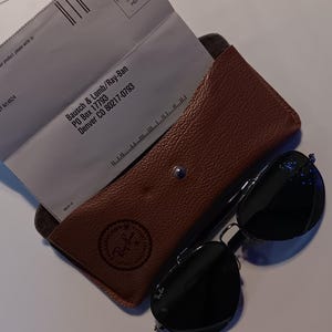 May include: A brown leather eyeglass case with a snap closure, containing two white envelopes. Black sunglasses with dark lenses rest beside the case. The Ray-Ban logo is visible on the case.