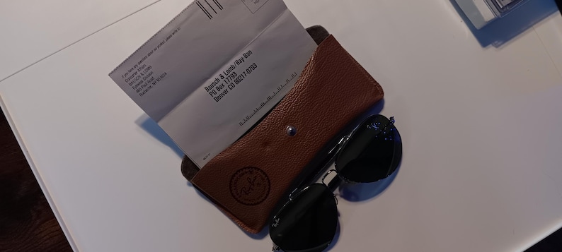 May include: A brown leather eyeglass case with a snap closure, containing two white envelopes. Black sunglasses with dark lenses rest beside the case. The Ray-Ban logo is visible on the case.